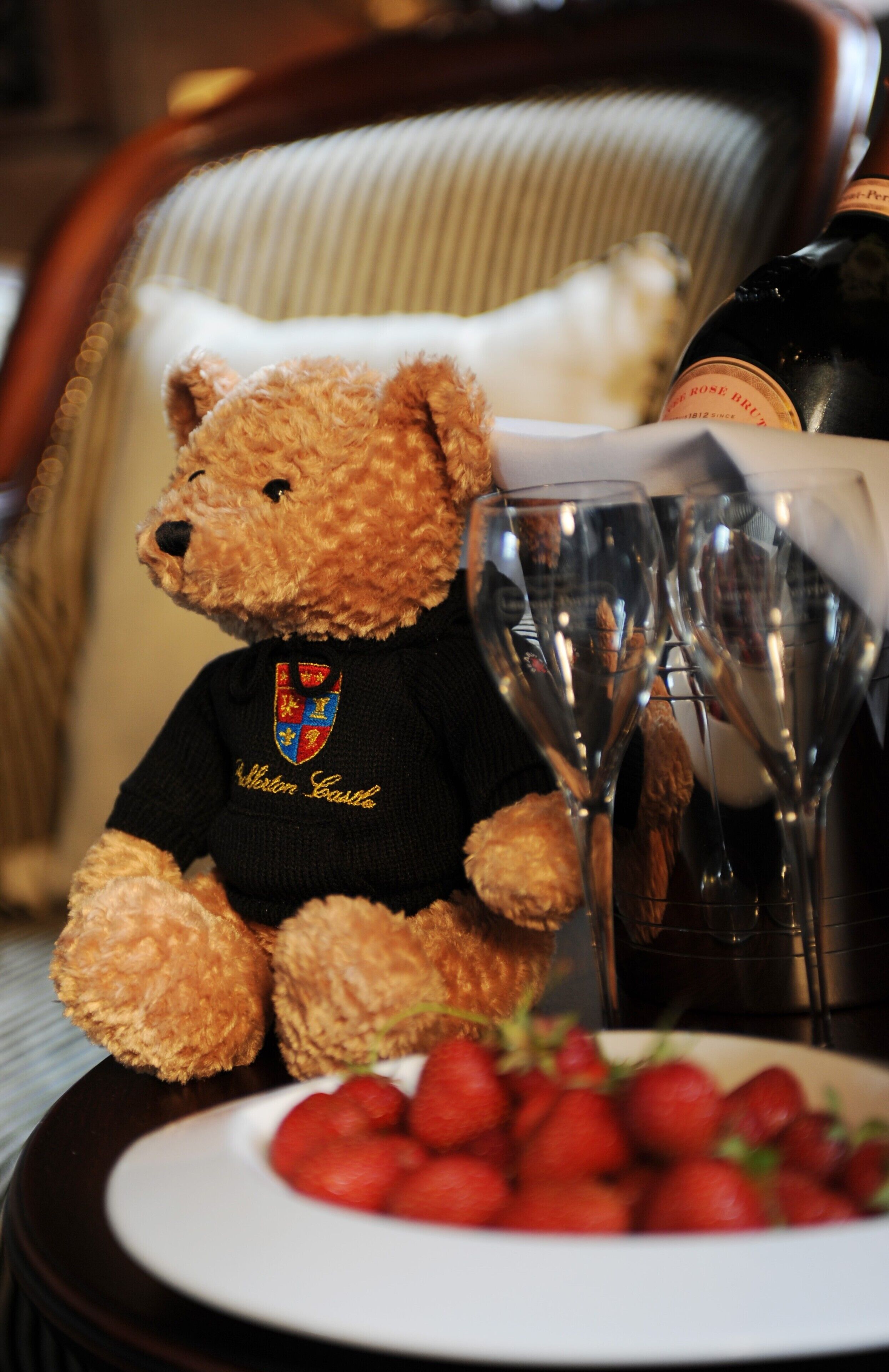 A teddy bear wearing a black shirt with a crest, sitting next to a bottle of wine and a glass.