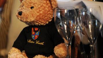 A teddy bear wearing a black shirt with a crest, sitting next to a bottle of wine and a glass.