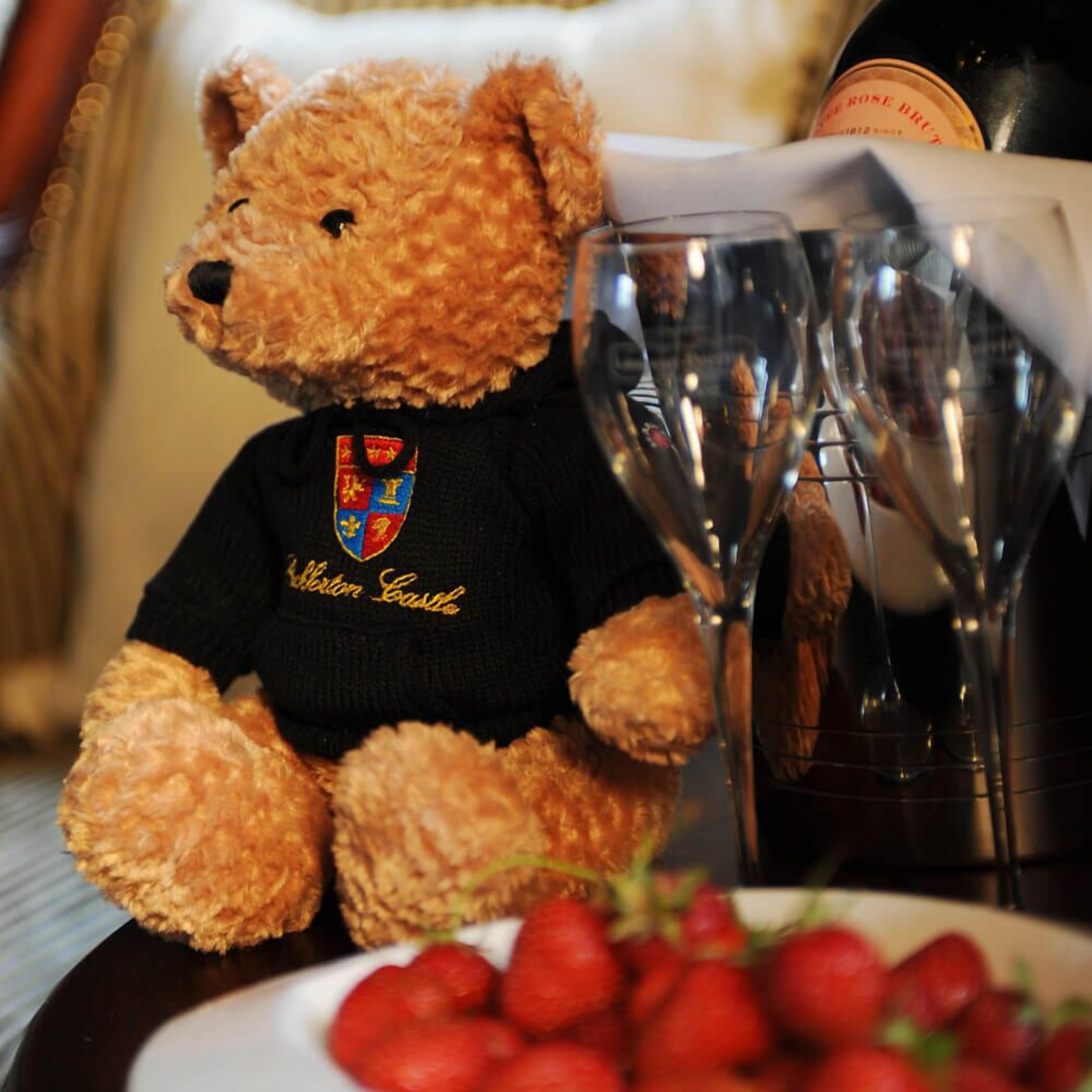 Un oso de peluche con una camisa negra que lleva un escudo, una botella de vino, dos copas de vino y un plato de fresas.