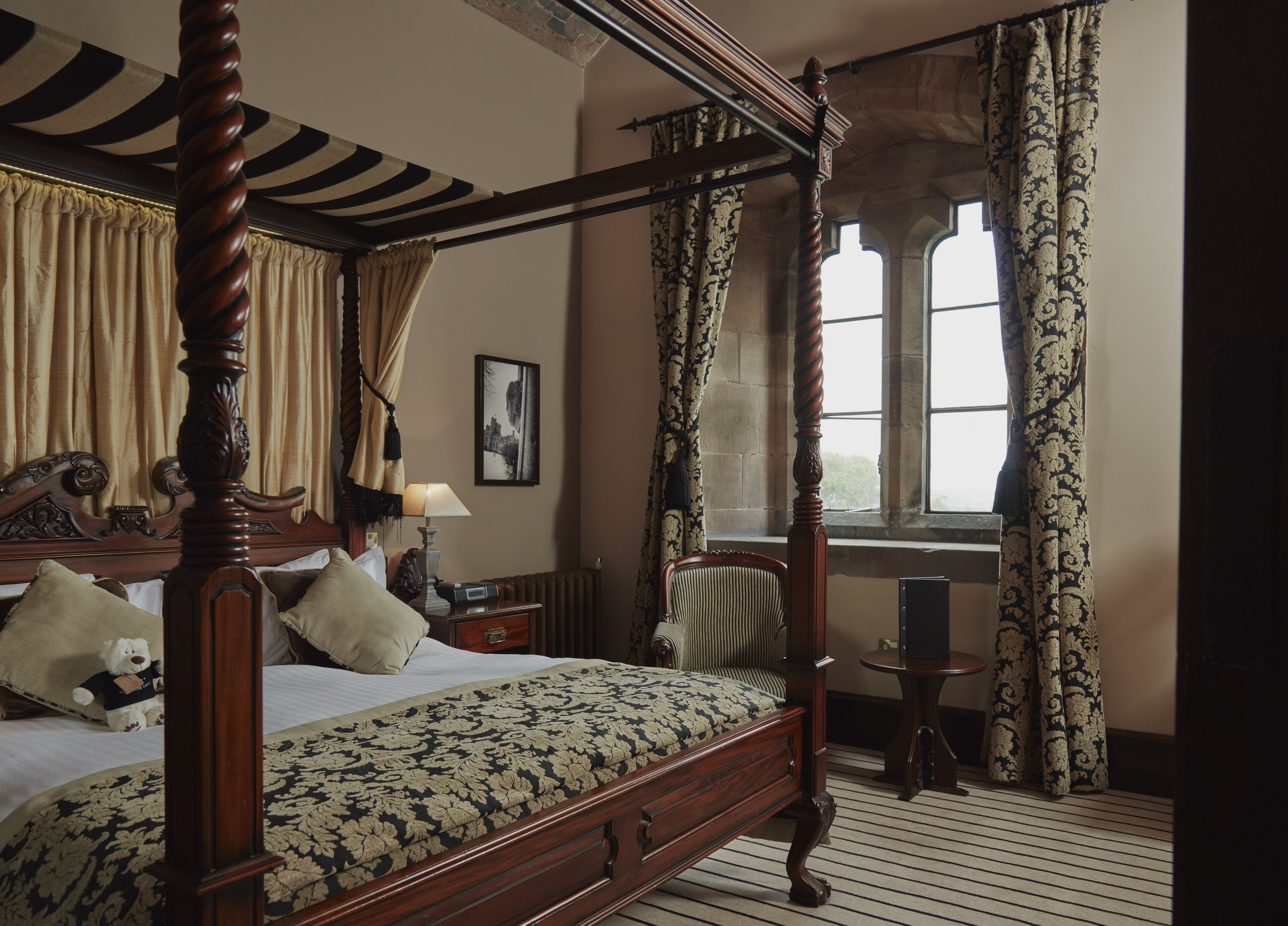 Una cama con dosel, un edredón floral, una mesita de noche con lámpara, una silla y una ventana con cortinas.