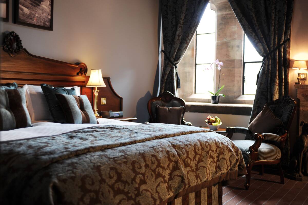 Une chambre d’hôtel avec un grand lit, une table de chevet, une chaise, une fenêtre avec des rideaux et une lampe.
