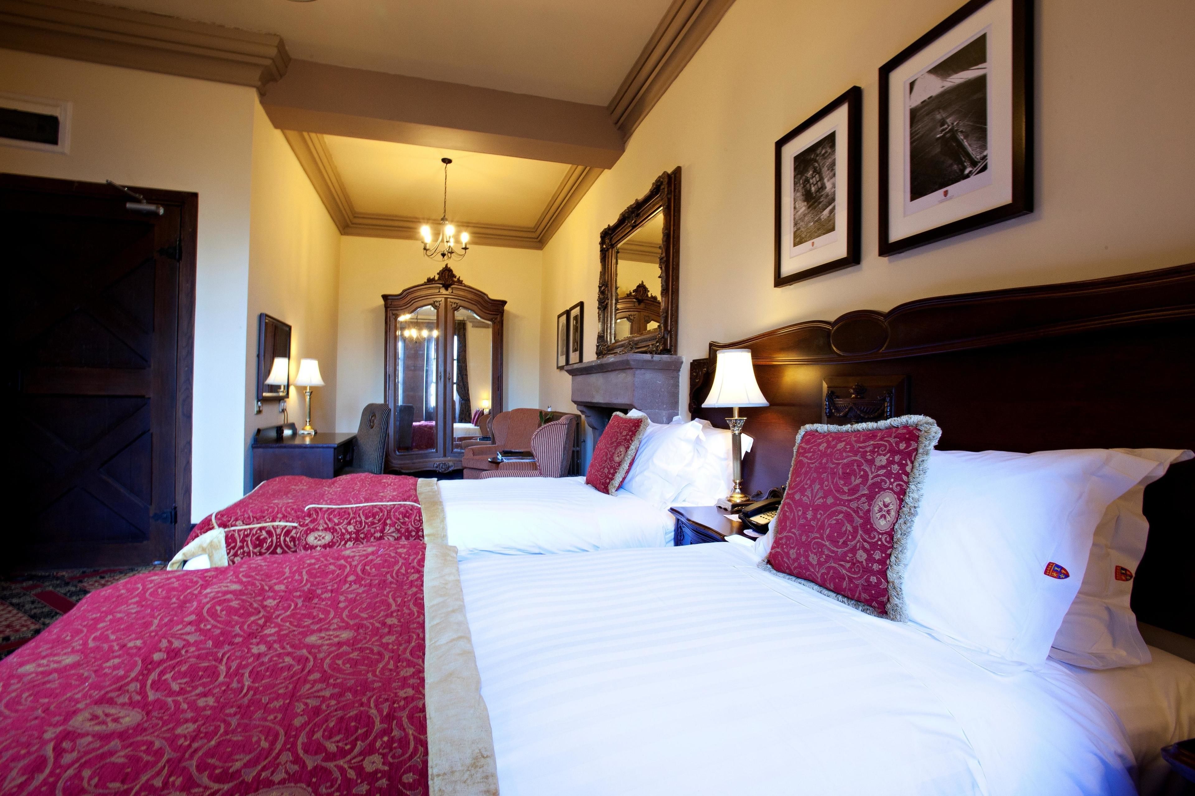 A hotel room with two beds, a wooden wardrobe, a desk with a lamp, and a mirror on the wall.