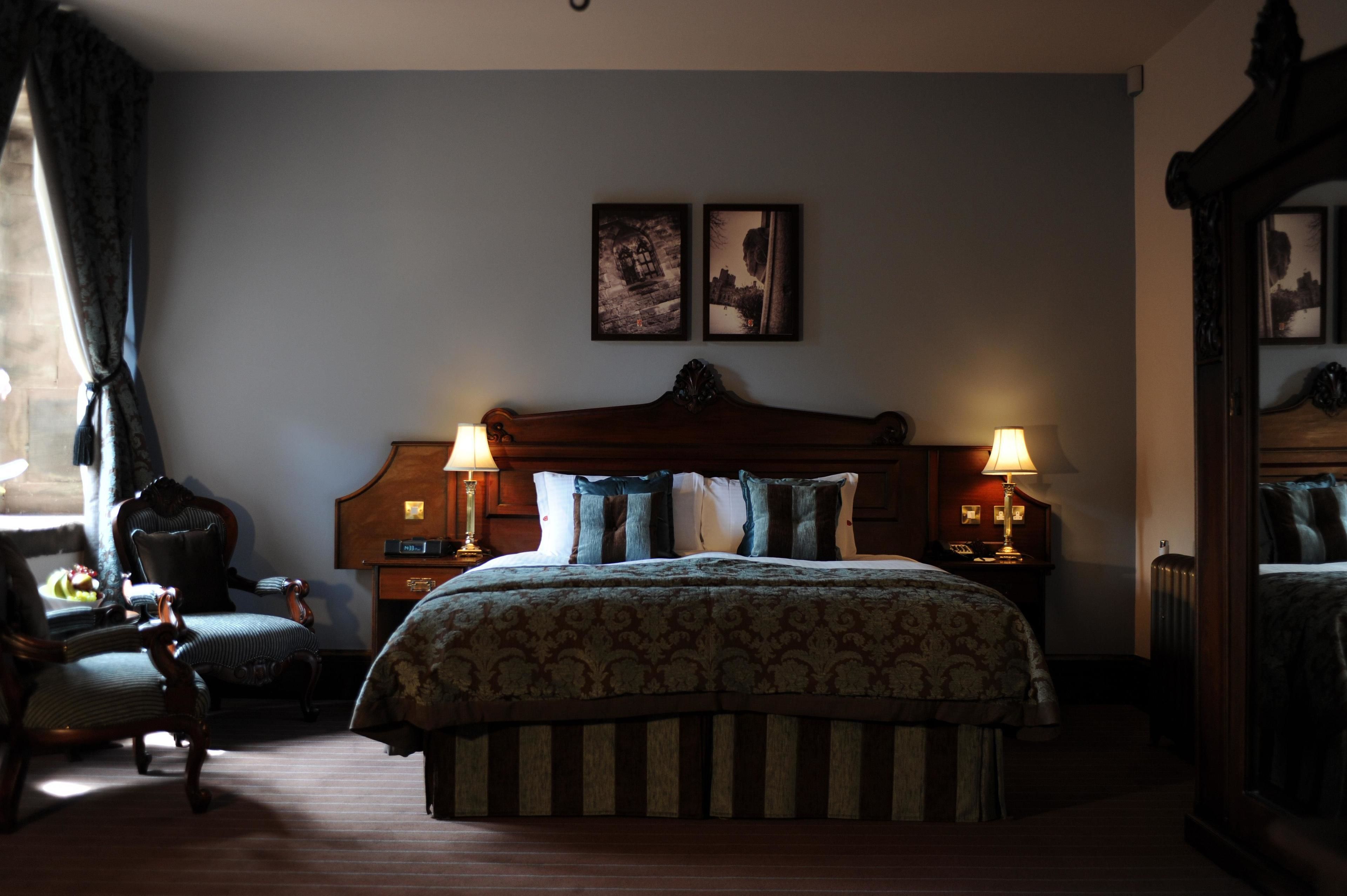 A hotel room with a large bed, two bedside lamps, a wooden headboard, a chair, and two framed pictures on the wall.