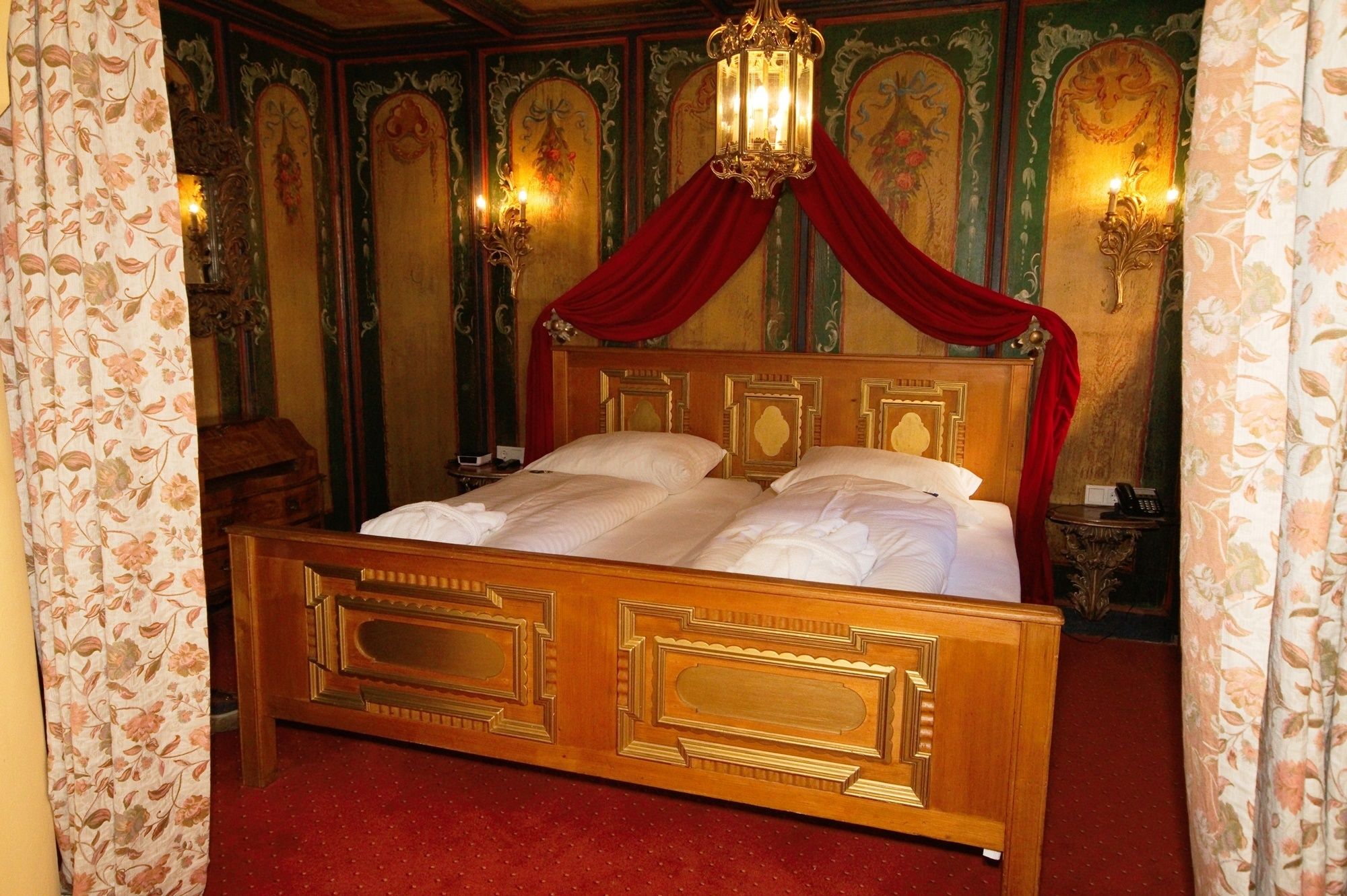 A four-poster bed with a red canopy in a room with ornate walls and floral curtains.