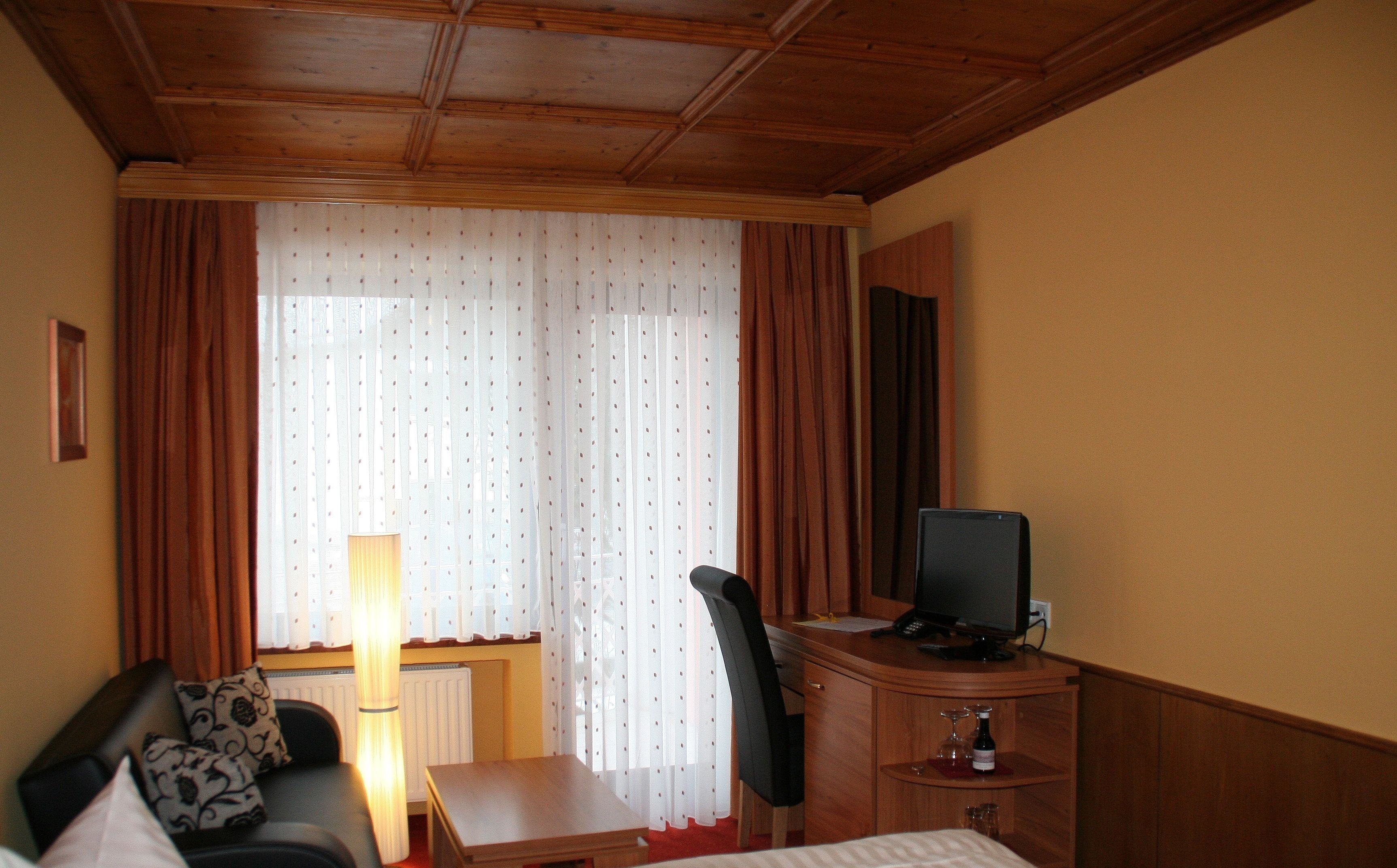A hotel room with a wooden ceiling, a sofa with patterned cushions, a desk with a television, and a window with curtains.