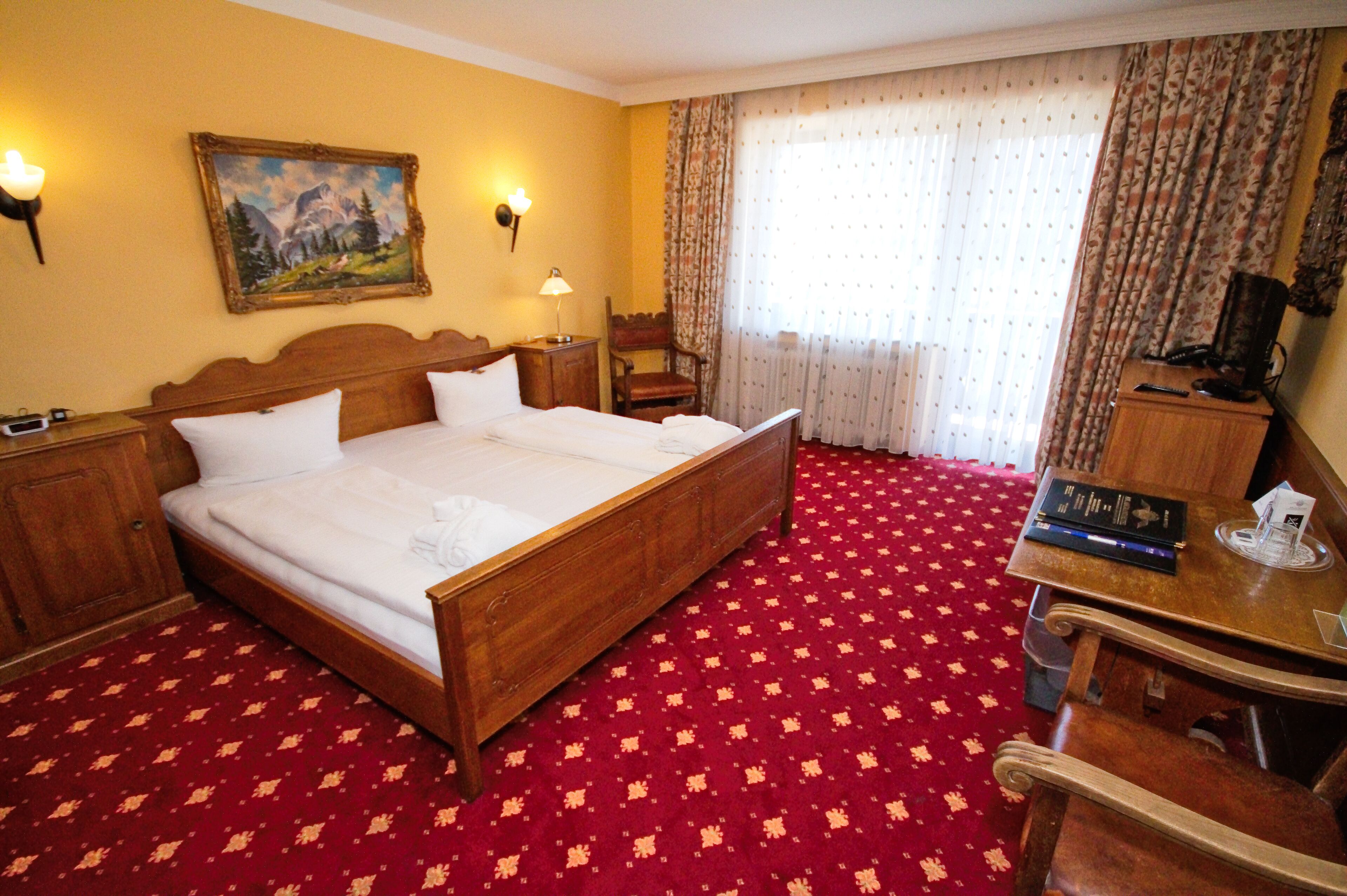 A hotel room with a wooden bed, a desk, a chair, a lamp, a painting, and a window with curtains.