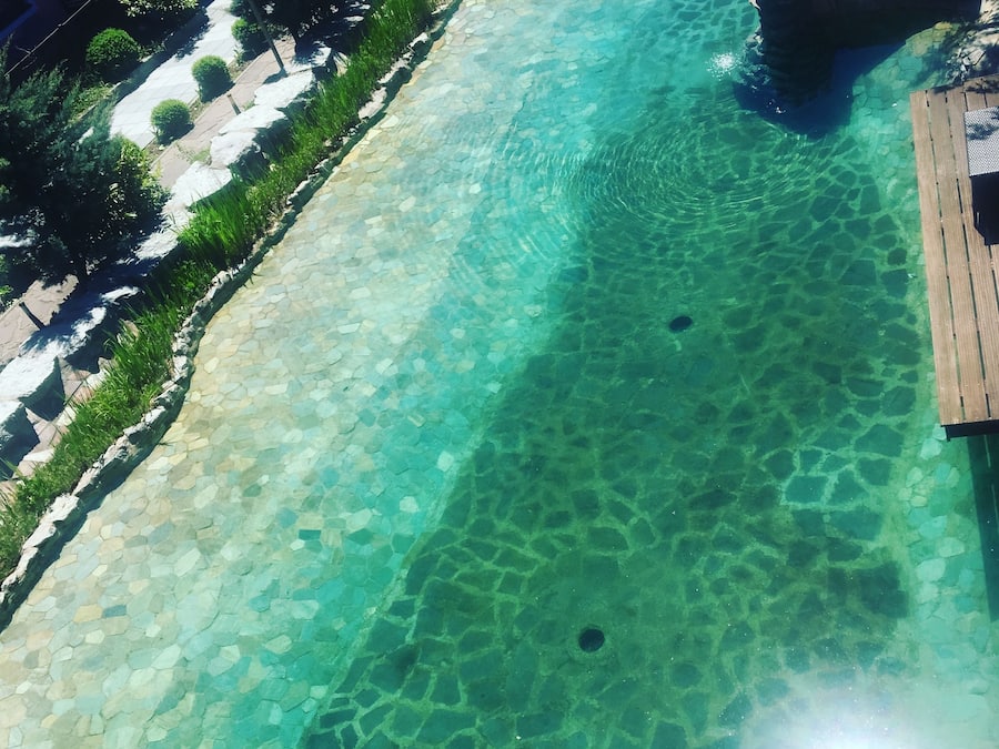 Piscina translúcida com borda de pedra, rodeada por vegetação e um deck de madeira.