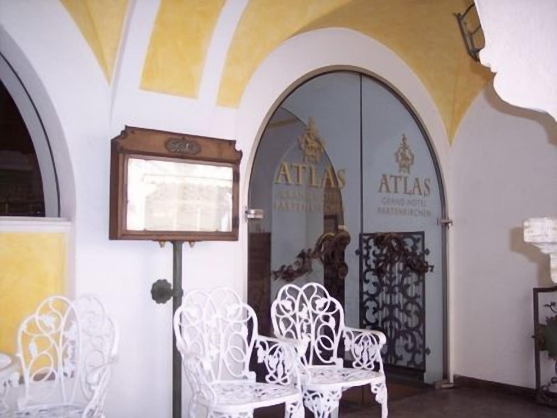 L’entrée d’un hôtel, avec un banc en métal blanc, une enseigne encadrée et une porte à deux battants portant l’inscription « ATLAS ».