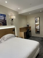 A hotel room with a bed, a desk, a chair, a mirror, and a wall-mounted lamp.