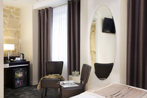 Ein Hotelzimmer mit Bett, Schreibtisch mit Lampe, Kühlschrank, Stuhl, Spiegel und Vorhängen.