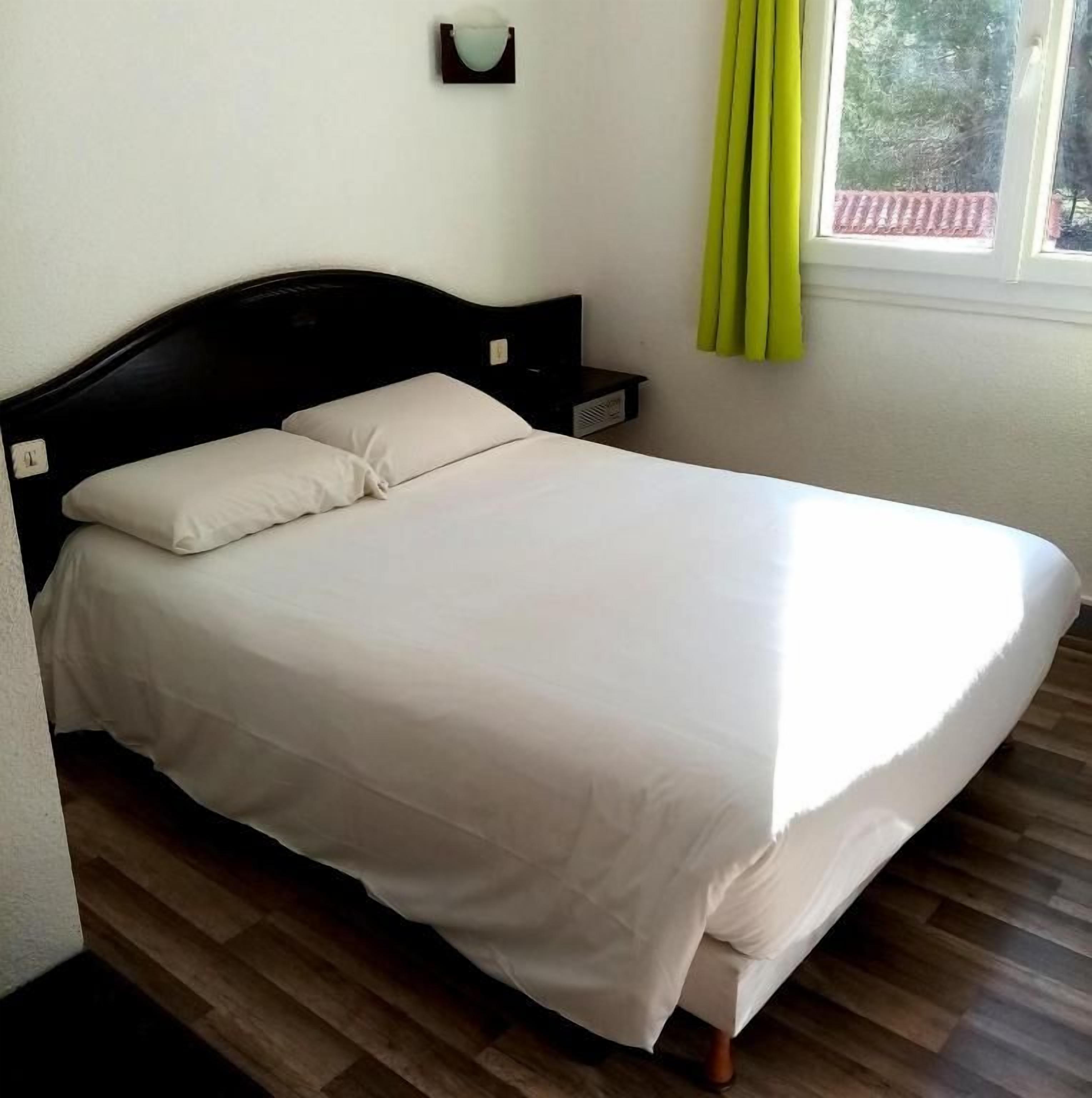 Un lit avec un matelas et des oreillers blancs, une tête de lit en bois, une table de chevet et une fenêtre avec des rideaux.