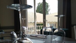 Une table à manger avec des verres à vin et des couverts, une vue sur un balcon aménagé avec du mobilier d’extérieur, et une porte donnant sur l’extérieur.