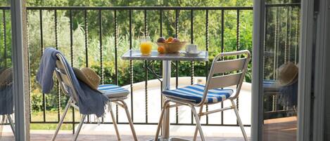 A balcony with two chairs, a table with a basket of fruit, a glass of orange juice, and a view of lush greenery.
