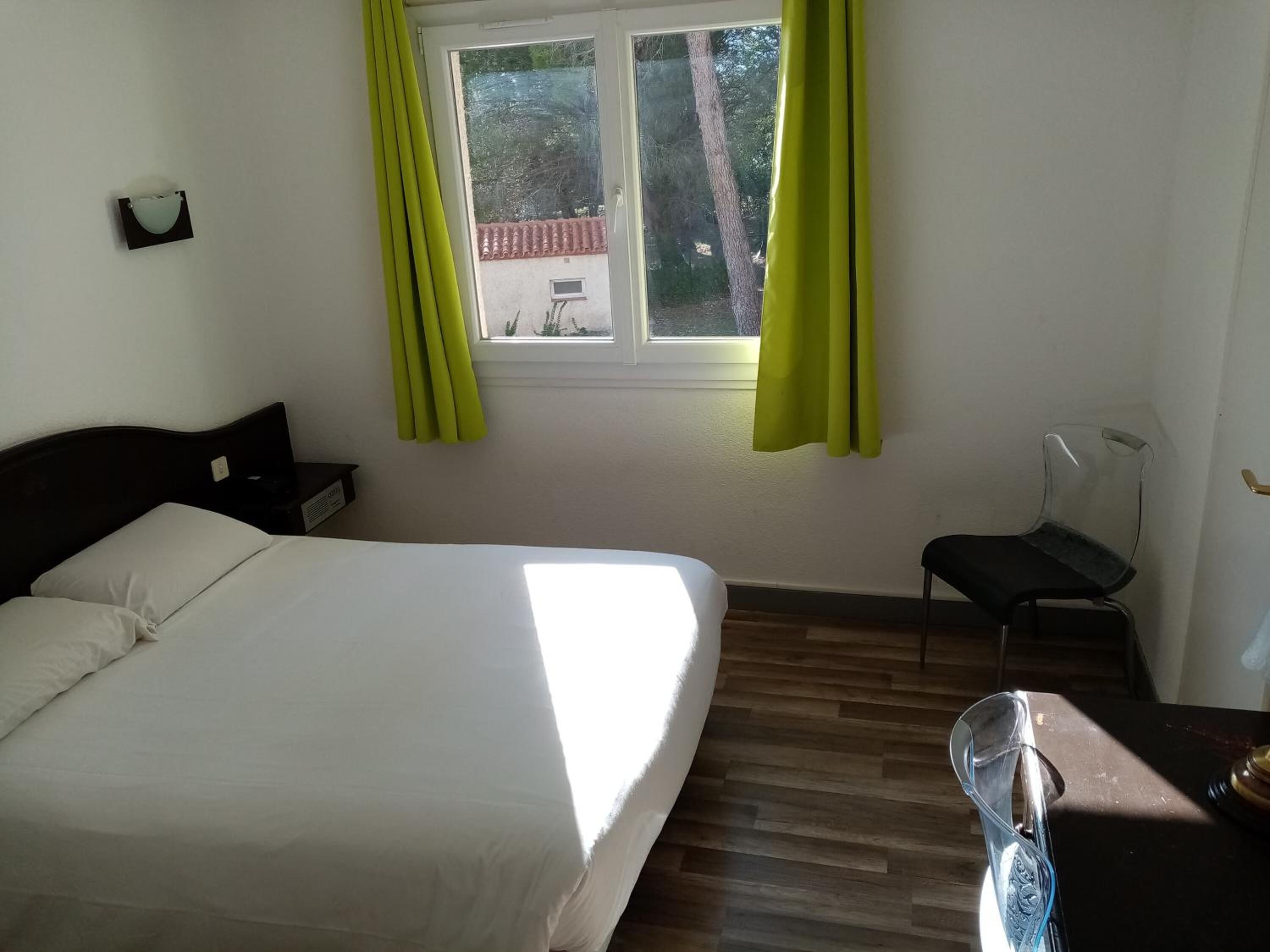 A bedroom with a bed, a chair, a desk, a window with curtains, and a view of trees and a building outside.