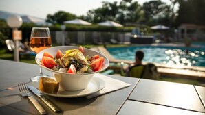 Une table à manger avec un saladier rempli de salade, un verre de vin, une fourchette et un couteau. Au fond, on aperçoit une piscine et une personne assise au bord de la piscine.