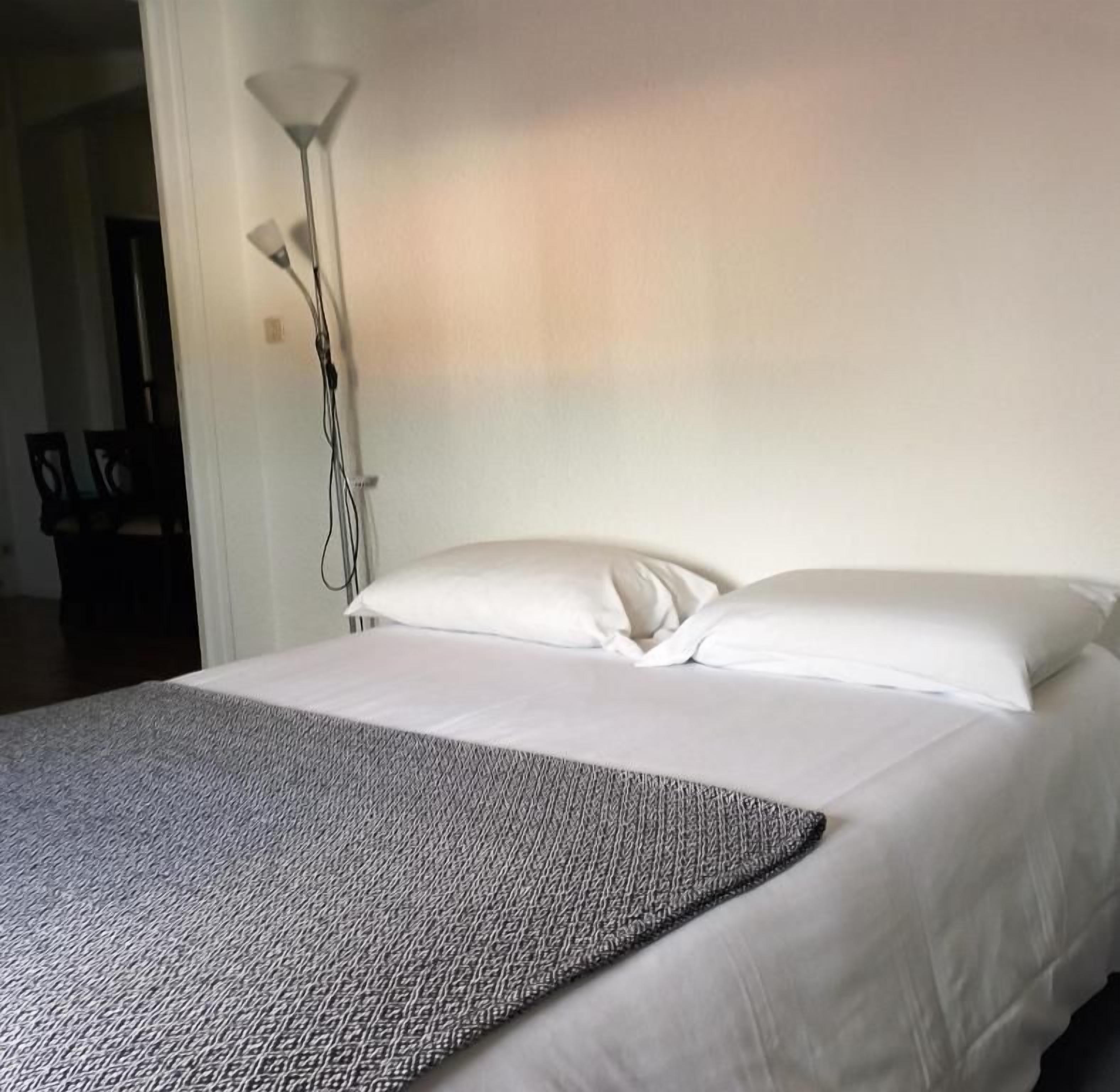 Une chambre d’hôtel avec un lit, un oreiller blanc, une couverture blanche et une lampe sur pied.