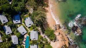 Luchtbeeld van een kustresort met meerdere bungalows, een centraal gelegen zwembad en een strand met rotsformaties.