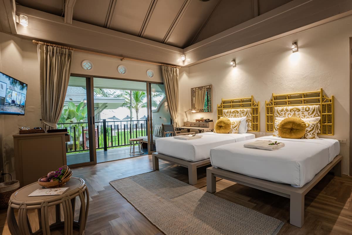 Une chambre d’hôtel avec deux lits, un bureau, une chaise et un balcon donnant sur des palmiers.