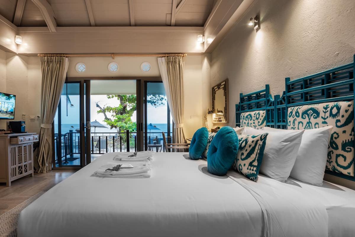 Une chambre d’hôtel avec un grand lit, un bureau équipé d’un ordinateur et une vue sur la mer, visible à travers une porte vitrée.