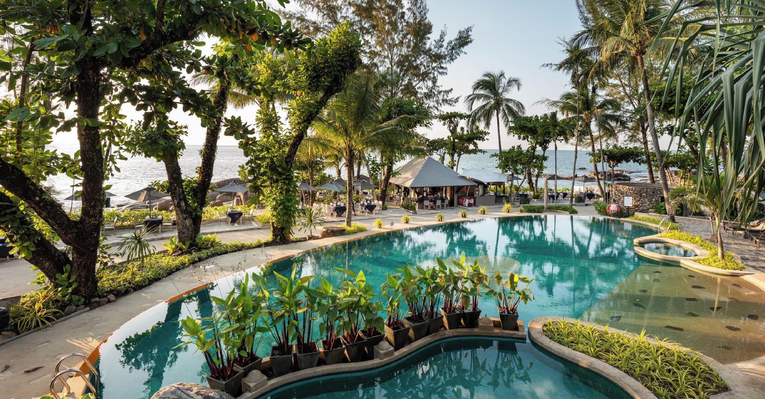A resort pool with a view of the sea, surrounded by palm trees and lounge areas.