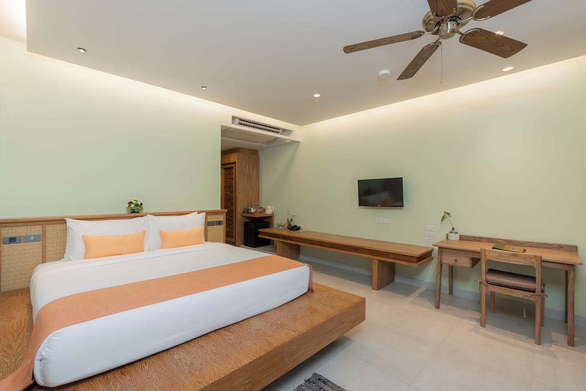 Un dormitorio con cama, escritorio, banca, ventilador de techo y televisor en la pared.