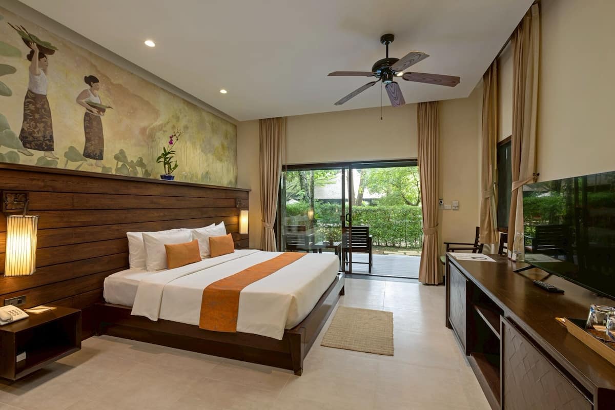 Habitación de hotel con una cama grande, ventilador de techo, televisión y balcón con vistas a zonas verdes.