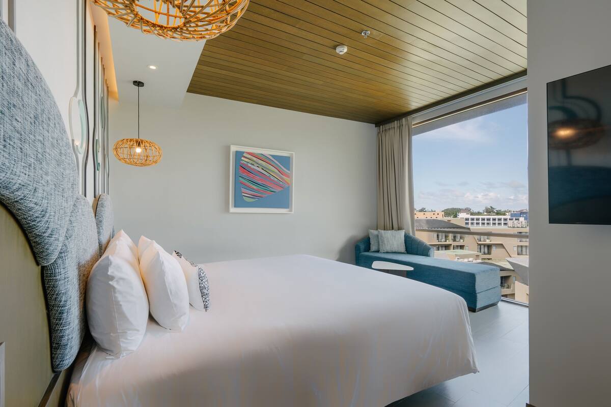 A modern hotel room with a large bed, a blue sofa, a city view, and a unique ceiling design.