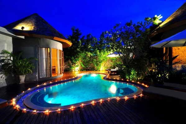 A swimming pool surrounded by wooden decking, with a curved edge and a small hot tub in the center.