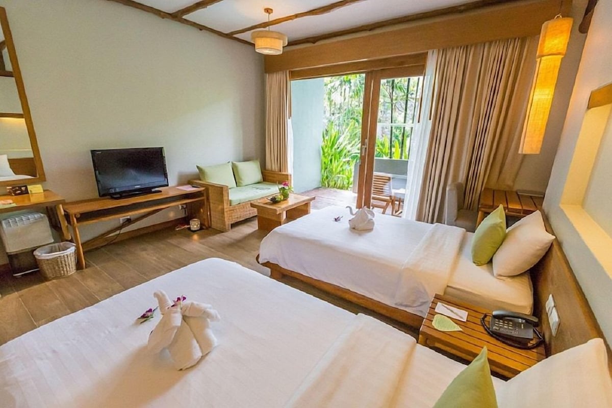 A hotel room with two beds, a television, a desk, and a view of greenery outside.