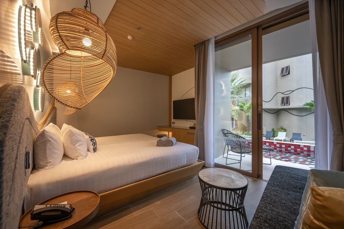 A modern hotel room with a bed, a small table, a TV, and a balcony with outdoor seating.