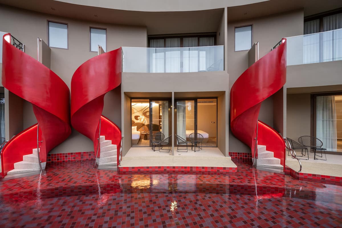 A modern hotel interior with a central pool area, red spiral staircases, and a seating area with chairs.