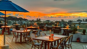 An outdoor dining area with tables and chairs, a large umbrella, and a view of the sunset over a cityscape. - Metadee Concept Hotel (Karon)