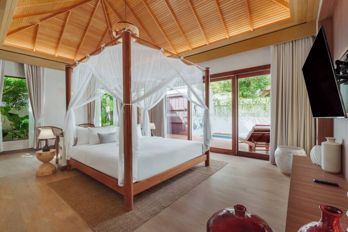 A spacious bedroom with a four-poster bed, a flat-screen TV, and a large window offering a view of the outdoors.