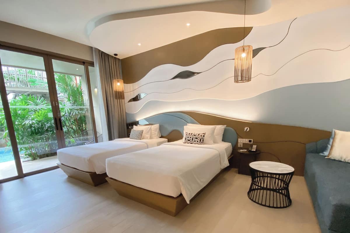 A modern hotel room with two beds, a sofa, a round table, and a view of greenery through large windows.
