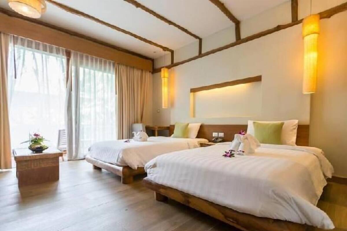 A hotel room with two beds, wooden flooring, a nightstand, a lamp, and a window with curtains.
