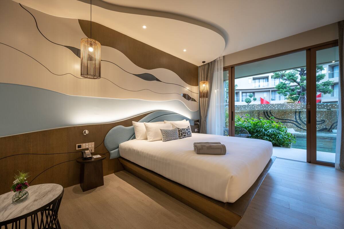 A modern hotel room with a large bed, a small table with a vase, and a view of the outdoors through a glass door.