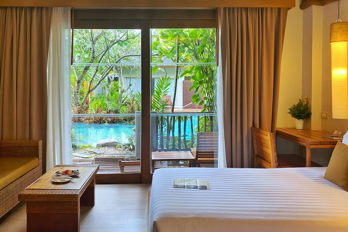 A hotel room with a bed, a wooden bench, a table with a plate and a bowl, and a view of a swimming pool and greenery through large windows.