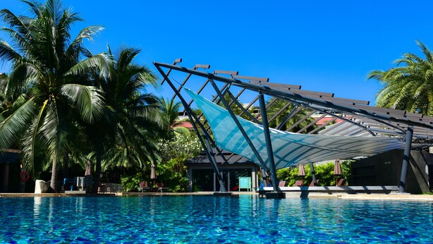 Una piscina azul cristalina con una zona sombreada, palmeras y un edificio al fondo.