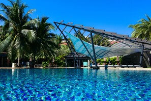 A clear blue pool with a shaded area, palm trees, and a building in the background. - R Mar Resort and Spa (Patong)