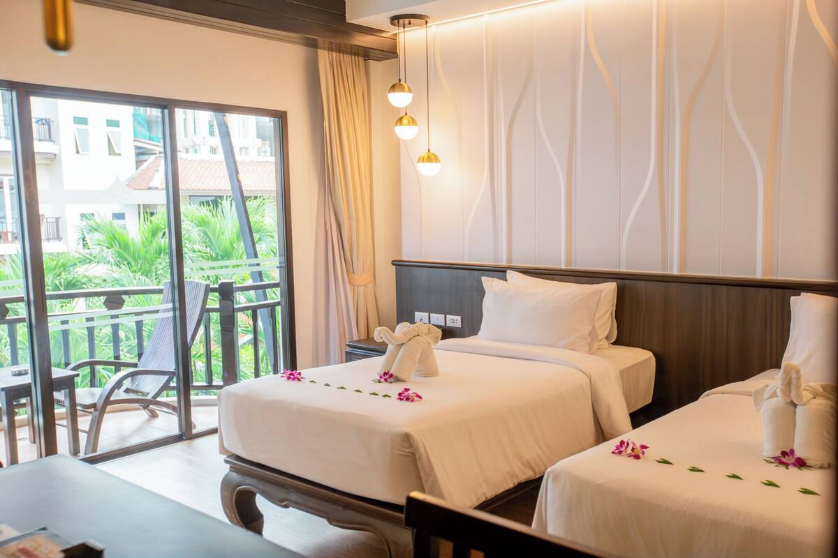 Quarto de hotel com duas camas, uma varanda com vista para edifícios e vegetação, e uma luminária moderna.