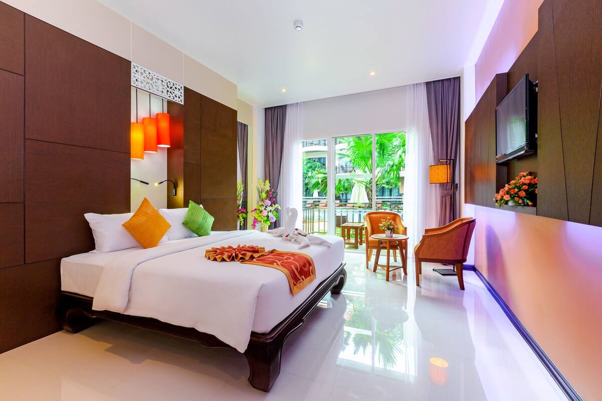 Quarto de hotel com uma cama grande, área de estar com uma poltrona e mesa, televisão fixada na parede e varanda com vista para o verde.