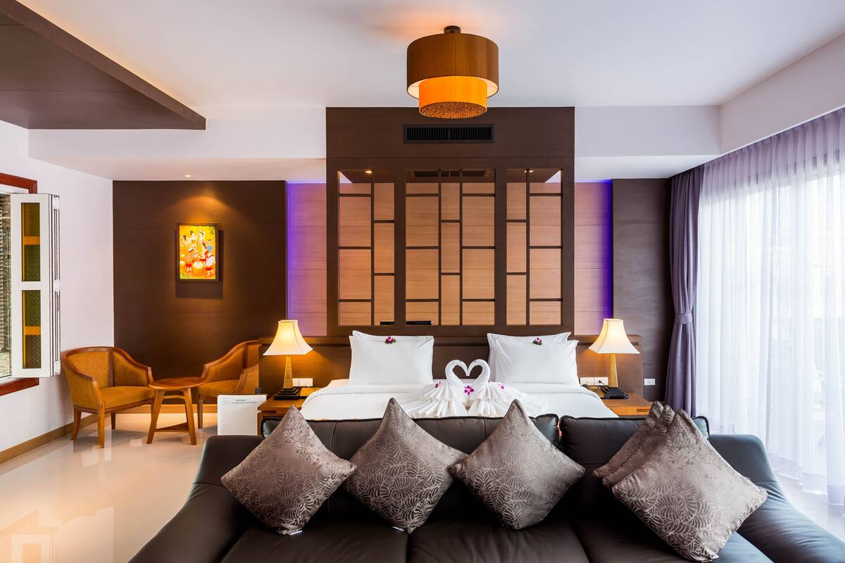 Quarto de hotel moderno com uma cama grande, duas mesas de cabeceira, um sofá e uma janela ampla com cortinas.