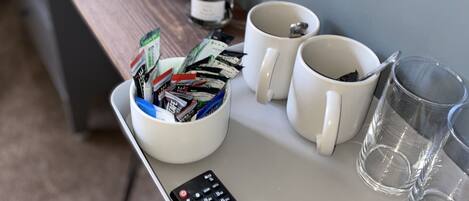 A tray with a remote control, two white mugs, a glass, a potted plant, and a container of snacks.