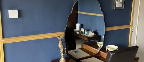 A room with a desk, chair, and wall-mounted mirror. There are framed pictures on the wall and a small trash bin on the floor.