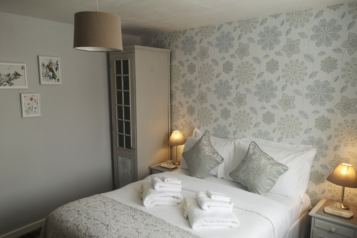 A neatly made bed with white linens and pillows, a bedside table with a lamp, and floral wallpaper.