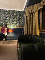 Una habitación con pared azul oscuro, papel tapiz estampado, sofá de cuero verde, una mesita redonda con lámpara y una ventana con cortinas estampadas.