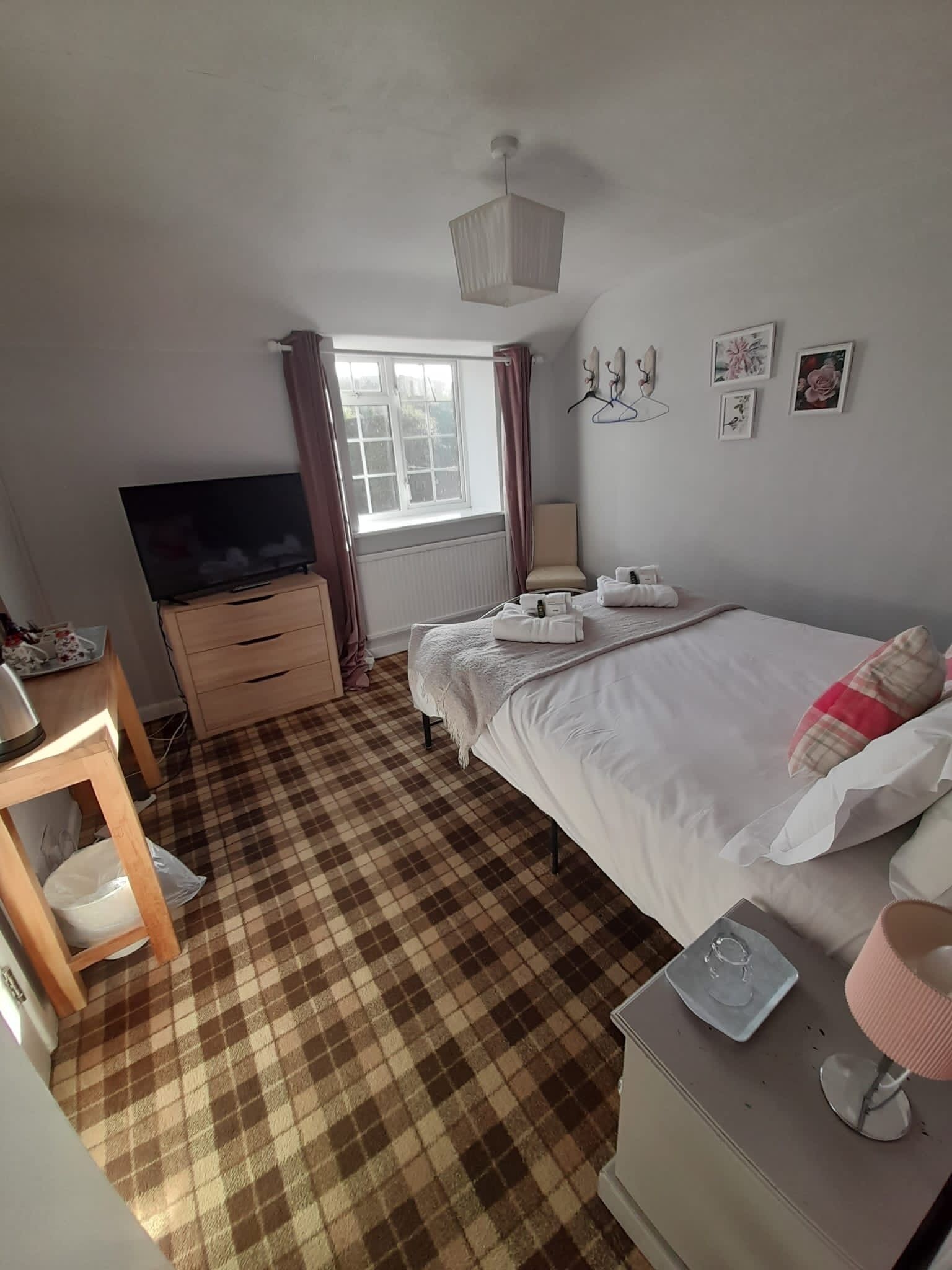 Una habitación de hotel con piso a cuadros, una cama con sábanas blancas, un televisor sobre un aparador de madera, una ventana con cortinas y una pared con cuadros enmarcados.