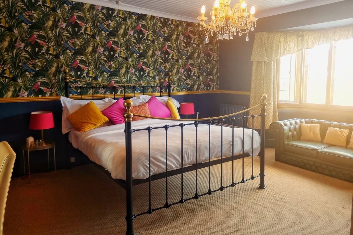 A bedroom with a bed, a chandelier, a patterned wall, a window, and a sofa.