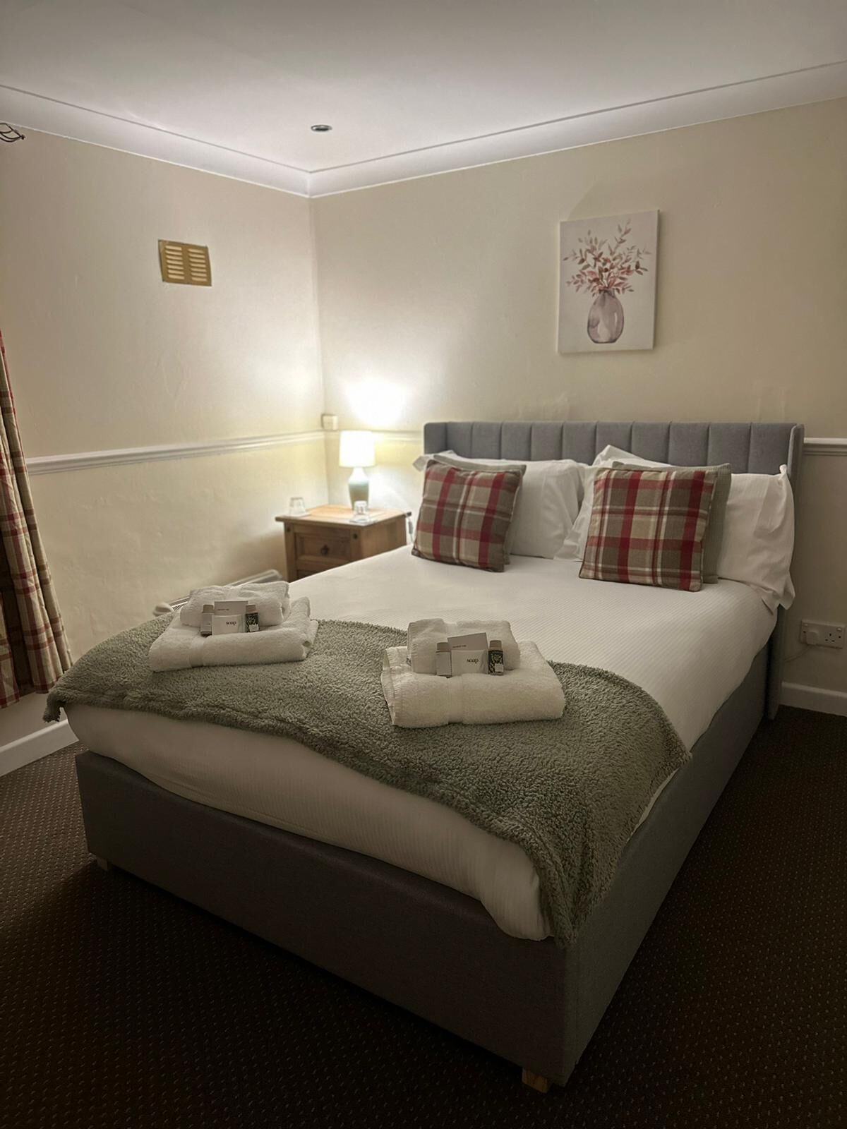 Een net opgemaakt bed met een grijs hoofdeinde, witte lakens en geruite kussens. Een groene plaid is over de voetteinde van het bed gedrapeerd. Een klein nachtkastje met een lamp en een opgevouwen handdoek is zichtbaar.