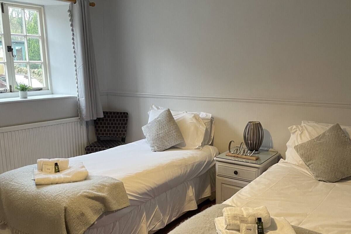 A hotel room with two beds, a window with curtains, a bedside table with a lamp, and a radiator.